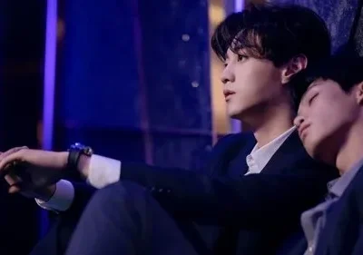 Dois homens jovens em ternos sob luz azul. Um olha para cima, o outro dorme em seu ombro. Clima íntimo e melancólico.