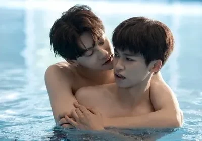 Dois jovens sem camisa se abraçam numa piscina. Um sussurra no ouvido do outro, que tem uma expressão surpresa e intensa.