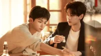 Dois jovens asiáticos à mesa. Um de suéter olha para baixo, outro de blazer o observa segurando um copo. Luz quente e clima íntimo.