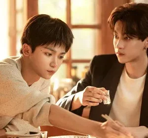 Dois jovens asiáticos à mesa. Um de suéter olha para baixo, outro de blazer o observa segurando um copo. Luz quente e clima íntimo.