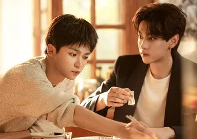 Dois jovens asiáticos à mesa. Um de suéter olha para baixo, outro de blazer o observa segurando um copo. Luz quente e clima íntimo.