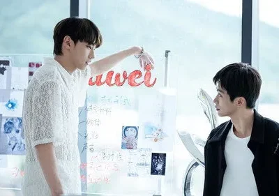 Dois jovens em um escritório moderno. Um aponta para um quadro de vidro cheio de ideias; o outro o observa. Clima colaborativo.