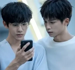 Dois jovens, um de óculos e top azul claro, segura um celular. O outro, em suéter branco, o observa com braço no ombro. Clima íntimo e curioso.