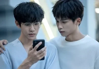 Dois jovens, um de óculos e top azul claro, segura um celular. O outro, em suéter branco, o observa com braço no ombro. Clima íntimo e curioso.