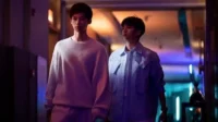 Dois jovens caminham por um corredor escuro com luzes neon roxas e azuis. Um de moletom cinza, outro de jaqueta listrada. Clima misterioso.