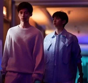 Dois jovens caminham por um corredor escuro com luzes neon roxas e azuis. Um de moletom cinza, outro de jaqueta listrada. Clima misterioso.
