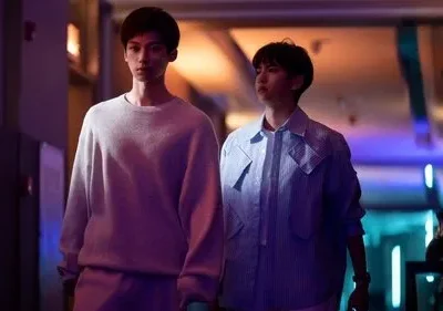 Dois jovens caminham por um corredor escuro com luzes neon roxas e azuis. Um de moletom cinza, outro de jaqueta listrada. Clima misterioso.