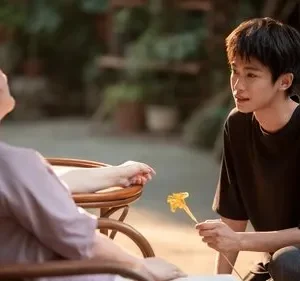 Jovem ajoelhado oferece flor amarela a mulher em cadeira. Cenário externo com luz dourada, expressa ternura e cuidado.