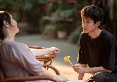 Jovem ajoelhado oferece flor amarela a mulher em cadeira. Cenário externo com luz dourada, expressa ternura e cuidado.