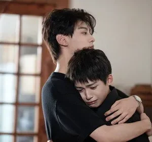 Dois jovens em um abraço apertado; um chora no ombro do outro. Fundo de interior com porta de madeira e banner amarelo.