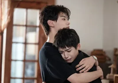 Dois jovens em um abraço apertado; um chora no ombro do outro. Fundo de interior com porta de madeira e banner amarelo.