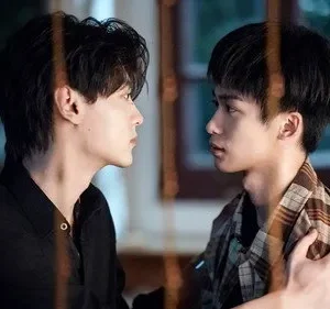 Dois homens jovens se encaram de perfil em um ambiente com pouca luz. Um, de xadrez, toca o ombro do outro, de preto. Clima íntimo e tenso.