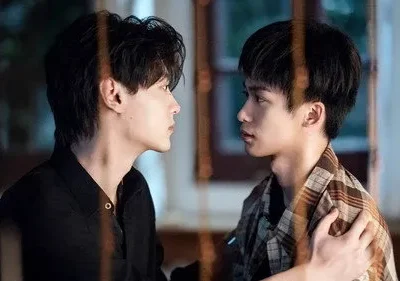 Dois homens jovens se encaram de perfil em um ambiente com pouca luz. Um, de xadrez, toca o ombro do outro, de preto. Clima íntimo e tenso.