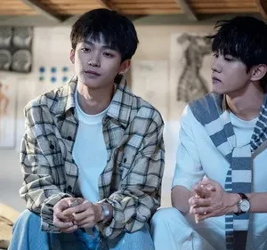 Dois jovens sentados em quarto com diagramas médicos. Um de camisa xadrez, outro de branco com tecido listrado. Clima sereno.