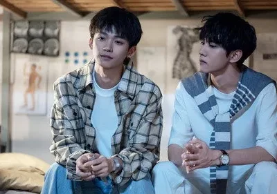 Dois jovens sentados em quarto com diagramas médicos. Um de camisa xadrez, outro de branco com tecido listrado. Clima sereno.