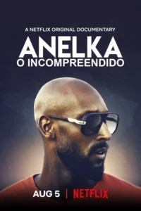 Homem careca e barbudo de pele escura, com óculos escuros e camisa laranja, olhando para a direita contra fundo escuro.