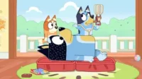 Um cão adulto deitado, com um filhote azul o ordenando com um brinquedo. Outro filhote laranja observa. Cena doméstica alegre e ensolarada.