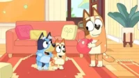 Cão adulto laranja sorri segurando balão vermelho. Dois filhotes, um azul e outro laranja, olham surpresos em sala aconchegante.