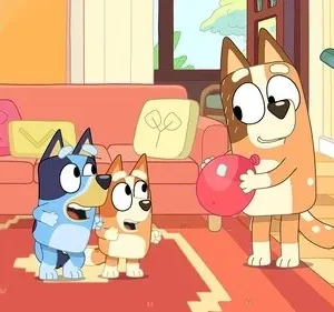 Cão adulto laranja sorri segurando balão vermelho. Dois filhotes, um azul e outro laranja, olham surpresos em sala aconchegante.