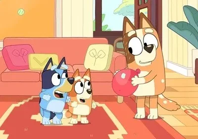 Cão adulto laranja sorri segurando balão vermelho. Dois filhotes, um azul e outro laranja, olham surpresos em sala aconchegante.