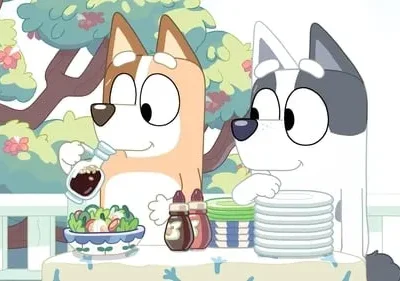Dois cães animados preparam uma refeição ao ar livre. Um cão veste salada, enquanto o outro organiza pratos. Clima feliz.