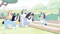 Família de cães estilizados brinca de trenzinho em jardim ensolarado. Dois cães grandes carregam dois pequenos, enquanto um terceiro lidera com guia.