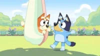 Cachorro laranja brinca numa rede com cachorro azul sorrindo em um parque. Cena alegre e divertida sob o sol.