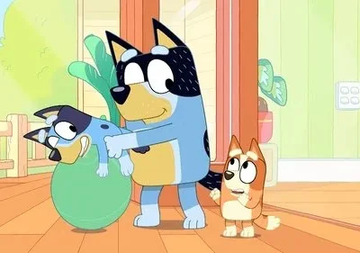 Cão azul adulto segura filhote azul brincalhão sobre bola verde. Um filhote laranja observa surpreso. Cenário doméstico alegre.