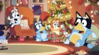 Quatro cães animados, dois adultos e dois filhotes, em uma sala de Natal caótica. Presentes voam, todos parecem surpresos e animados.