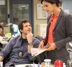 Homem sorridente com distintivo olha para mulher de terno examinando papéis em um escritório movimentado. Clima leve e profissional.