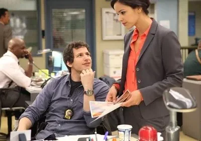 Homem sorridente com distintivo olha para mulher de terno examinando papéis em um escritório movimentado. Clima leve e profissional.