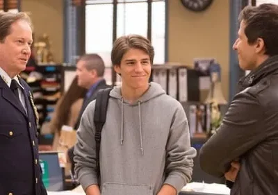 Três homens conversam em um escritório. Um policial, um jovem sorridente de moletom e um homem de jaqueta. Clima amigável.