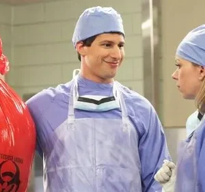 Dois cirurgiões em uniforme azul. Um homem sorri maliciosamente segurando um saco de lixo vermelho de risco biológico. Uma mulher o olha séria.