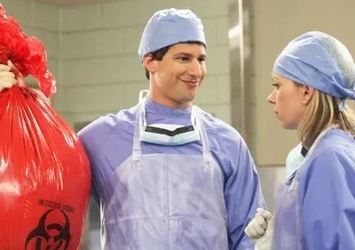 Dois cirurgiões em uniforme azul. Um homem sorri maliciosamente segurando um saco de lixo vermelho de risco biológico. Uma mulher o olha séria.