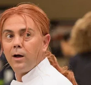 Um chef ruivo de jaleco branco, com expressão de surpresa, olhos arregalados e vesgos. Fundo desfocado.