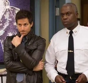 Um detetive jovem, pensativo em jaqueta de couro, ao lado de um oficial mais velho e sério em uniforme policial. Fundo de delegacia.