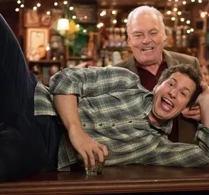 Jovem deitado em balcão de bar, gargalhando, com homem mais velho sorridente por trás. Ambiente animado com garrafas.