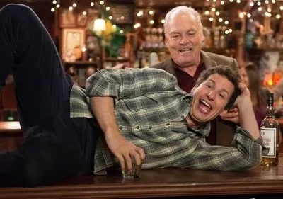 Jovem deitado em balcão de bar, gargalhando, com homem mais velho sorridente por trás. Ambiente animado com garrafas.