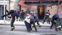 Bombeiros e civis em confronto/resgate intenso numa rua urbana. Um bombeiro carrega um homem, outros se agarram. Prédio danificado ao fundo.