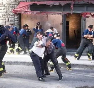 Bombeiros e civis em confronto/resgate intenso numa rua urbana. Um bombeiro carrega um homem, outros se agarram. Prédio danificado ao fundo.