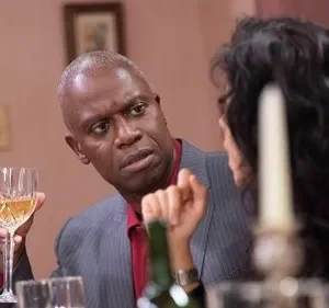 Homem de terno, com cabelo grisalho, segura taça de vinho e olha cético para pessoa à mesa num ambiente formal.