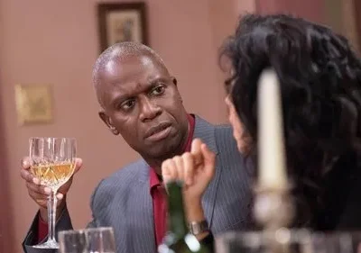 Homem de terno, com cabelo grisalho, segura taça de vinho e olha cético para pessoa à mesa num ambiente formal.