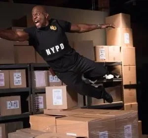 Policial musculoso, voando e atirando em armazém cheio de caixas. Jovem observa surpreso de um pilar. Cena de ação e aventura.