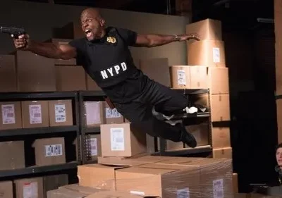 Policial musculoso, voando e atirando em armazém cheio de caixas. Jovem observa surpreso de um pilar. Cena de ação e aventura.