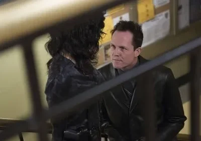 Homem de jaqueta escura olha sério para mulher de costas com cabelos longos. Cena interna, com grades em primeiro plano.