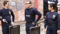Três policiais do NYPD (2 homens, 1 mulher) em um pátio de tijolos. Um homem sorri, um com colete POLICE e uma mulher séria.
