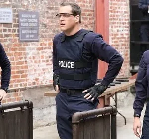 Três policiais do NYPD (2 homens, 1 mulher) em um pátio de tijolos. Um homem sorri, um com colete POLICE e uma mulher séria.