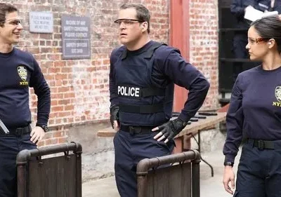 Três policiais do NYPD (2 homens, 1 mulher) em um pátio de tijolos. Um homem sorri, um com colete POLICE e uma mulher séria.