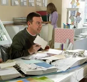 Três pessoas em uma loja de artesanato. Dois homens sentados à mesa, um gesticula com papel. Uma mulher os ouve atentamente.