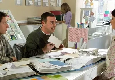 Três pessoas em uma loja de artesanato. Dois homens sentados à mesa, um gesticula com papel. Uma mulher os ouve atentamente.